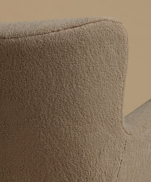 A close up image of an accent chair upholstered in textured taupe fabric with light natural wood legs, set against a solid warm beige background.