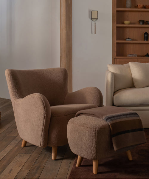 A cozy upholstered armchair in a warm taupe bouclé fabric sits beside a matching ottoman with a striped throw blanket, set in a styled living room with wood flooring and shelving.
