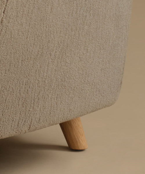 Close-up of a beige upholstered furniture corner with a light natural wood leg on a warm beige background.