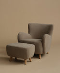 A modern upholstered armchair with a matching ottoman, both in a textured taupe fabric with light natural wood legs, set against a solid warm beige background.