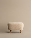 The Aspen Ottoman, a small, light beige piece with textured Sherpa fabric and wooden legs, sits against a plain beige background.