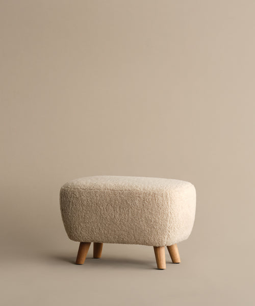 The Aspen Ottoman features a compact design, cream textured fabric, and four wooden legs, displayed on a plain beige background.
