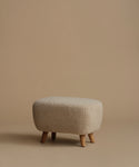 A low, square upholstered ottoman stool with a textured bouclé fabric in a light beige color and four tapered wooden legs, set against a solid warm beige background.
