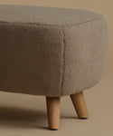 A close-up of a round upholstered ottoman  with a textured bouclé fabric in a warm taupe-beige color, supported by four tapered light wood legs, set against a solid beige background.