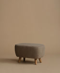 A low upholstered ottoman stool with a textured taupe fabric and four tapered wooden legs, set against a solid warm beige background.