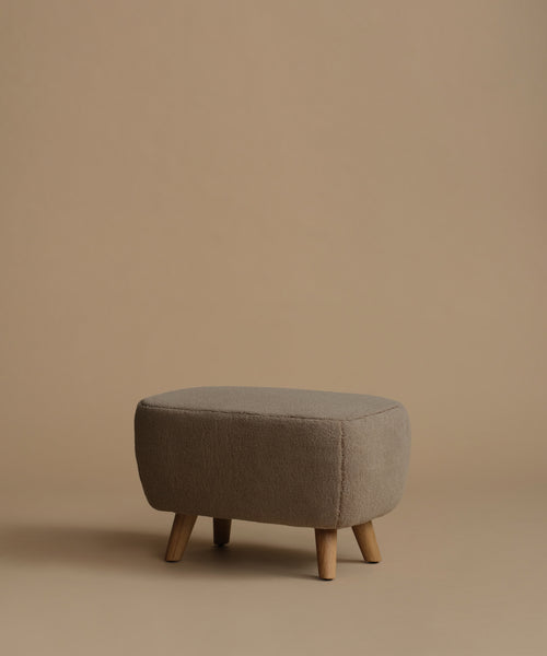 A low upholstered ottoman stool with a textured taupe fabric and four tapered wooden legs, set against a solid warm beige background.