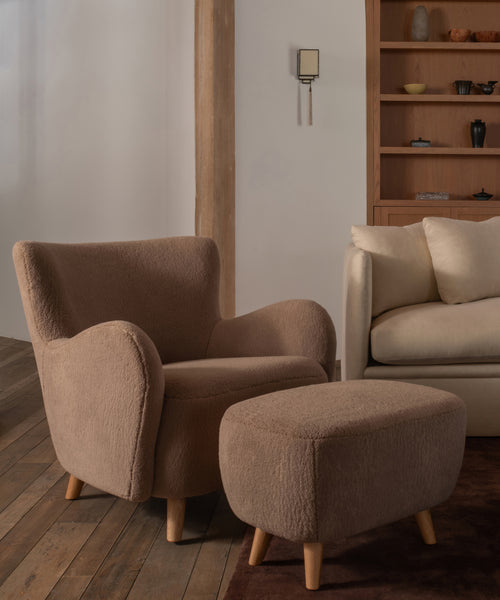 A low upholstered ottoman stool with a textured taupe fabric and four tapered wooden legs with a matching textured taupe fabric accent chair inside a home.