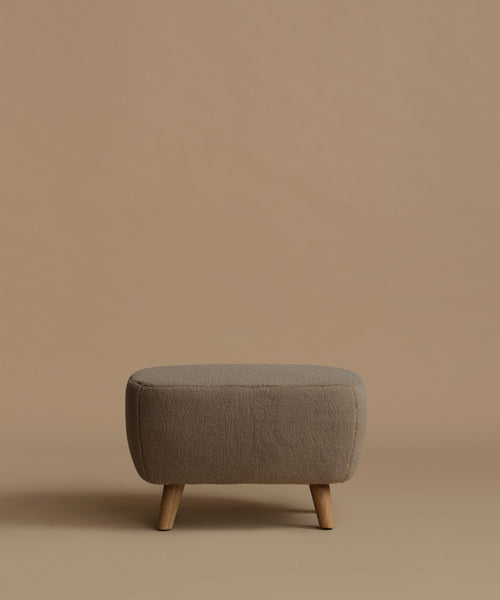 A low upholstered ottoman stool with a textured taupe fabric and four tapered wooden legs, set against a solid warm beige background.