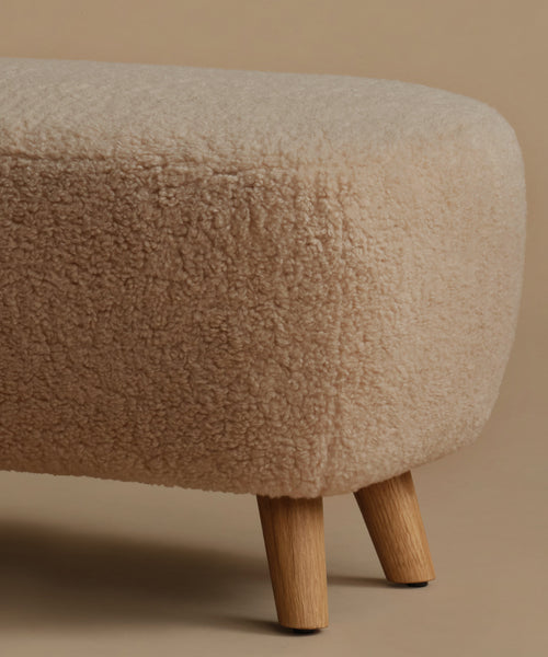 A low, square upholstered ottoman stool with a textured bouclé fabric in a light beige color and four tapered wooden legs, set against a solid warm beige background.