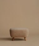 A low, square upholstered ottoman stool with a textured bouclé fabric in a light beige color and four tapered wooden legs, set against a solid warm beige background.