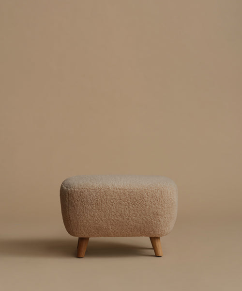A low, square upholstered ottoman stool with a textured bouclé fabric in a light beige color and four tapered wooden legs, set against a solid warm beige background.