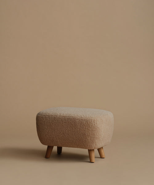 A low, square upholstered ottoman stool with a textured bouclé fabric in a light beige color and four tapered wooden legs, set against a solid warm beige background.