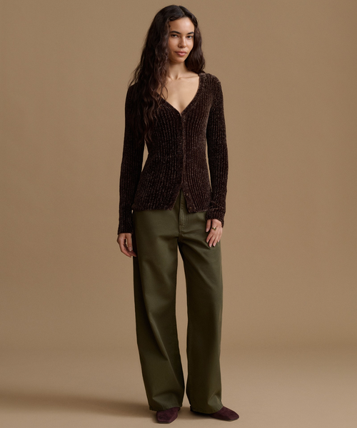 A woman stands against a plain warm beige studio background wearing a dark brown ribbed knit cardigan with a deep V-neck and olive green wide-leg trousers.