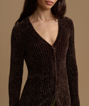 A close-up of a model wearing a dark brown ribbed knit cardigan with a deep V-neck and subtle shimmering texture, set against a solid warm beige background.