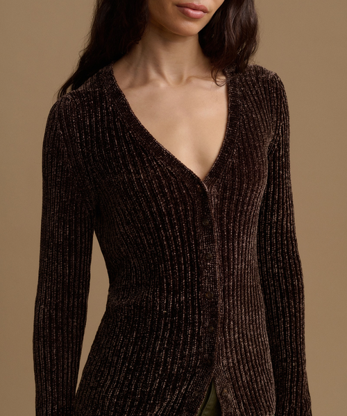 A close-up of a model wearing a dark brown ribbed knit cardigan with a deep V-neck and subtle shimmering texture, set against a solid warm beige background.