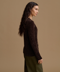 A woman in side profile wearing a dark brown ribbed knit sweater and olive green pants, photographed against a solid warm beige studio background.