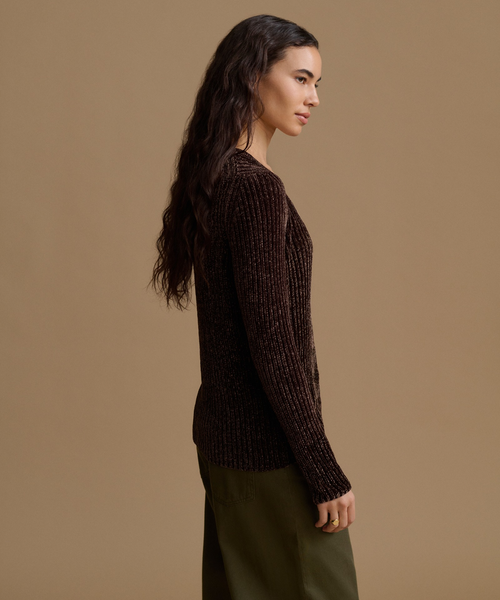 A woman in side profile wearing a dark brown ribbed knit sweater and olive green pants, photographed against a solid warm beige studio background.