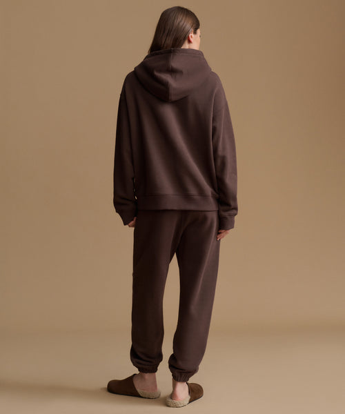 A model viewed from behind wearing a matching dark brown hoodie and jogger sweatpants set, standing against a solid beige studio background.