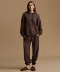A woman stands against a plain warm beige studio background wearing a matching dark brown hoodie and jogger sweatpants set, with brown slip-on slippers.