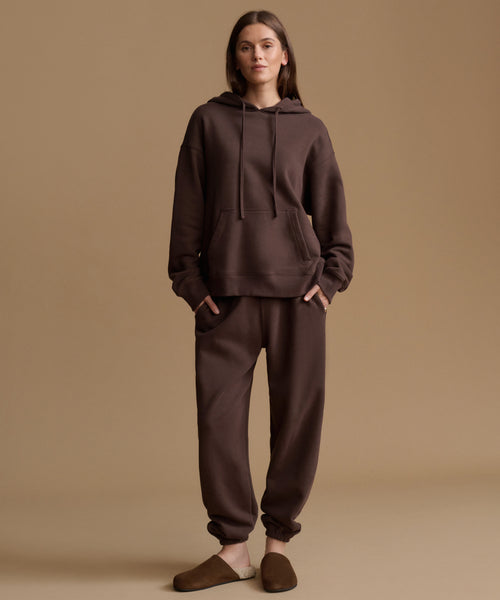A woman stands against a plain warm beige studio background wearing a matching dark brown hoodie and jogger sweatpants set, with brown slip-on slippers.