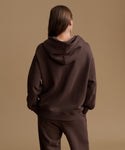 A model shown from the back wearing a dark brown oversized hoodie and matching dark brown sweatpants against a solid warm beige studio background.