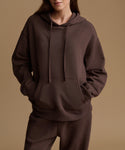 A model wearing a dark brown oversized pullover hoodie with matching sweatpants, shown from the neck down against a solid warm beige studio background.
