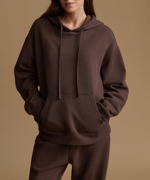 A model wearing a dark brown oversized pullover hoodie with matching sweatpants, shown from the neck down against a solid warm beige studio background.