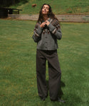 A woman stands on a grassy field wearing a gray knit cardigan with a brown and beige pattern, paired with dark wide-leg pants and black boots.