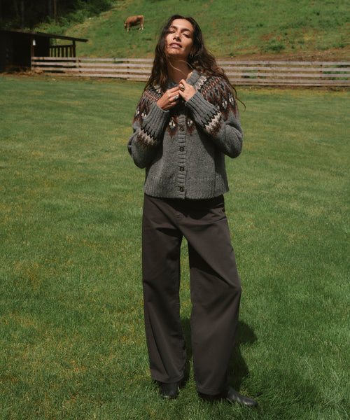 A woman stands on a grassy field wearing a gray knit cardigan with a brown and beige pattern, paired with dark wide-leg pants and black boots.