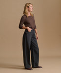A woman stands in profile wearing a fitted brown long-sleeve top and dark charcoal wide-leg pants against a plain warm beige studio background.