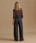 A woman is shown from behind wearing a dark brown long-sleeve top and dark charcoal gray wide-leg trousers, standing against a solid beige studio background.