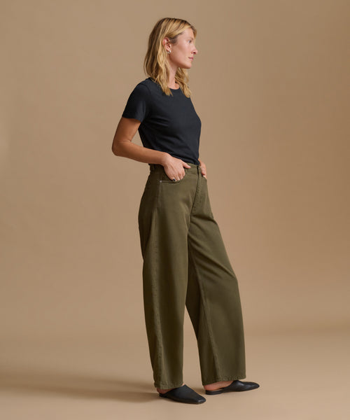 A woman stands in profile wearing olive green wide-leg trousers, a black short-sleeve t-shirt, and black flats against a solid beige studio background.