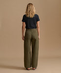 A woman viewed from behind wearing olive green wide-leg pants and a dark navy t-shirt, standing against a solid beige studio background.