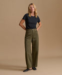 A woman stands against a plain beige studio background wearing olive green wide-leg trousers, a black short-sleeve top, and black flats.
