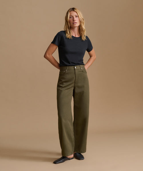 A woman stands against a plain beige studio background wearing olive green wide-leg trousers, a black short-sleeve top, and black flats.