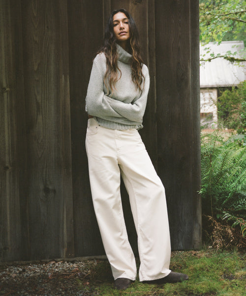 A woman stands outdoors against a dark wooden wall, wearing a light gray turtleneck sweater and off-white wide-leg trousers with dark shoes.