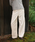 A person wearing off-white wide-leg pants and a light gray ribbed sweater, standing outdoors against a dark wooden fence with green plants and moss on the ground.