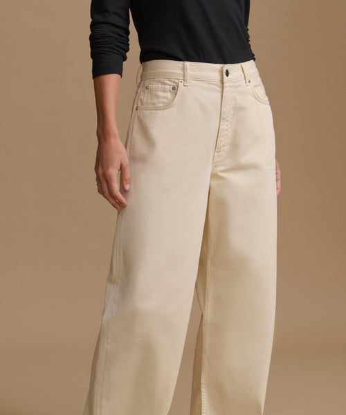 A model wearing high-waisted, wide-leg beige trousers and a black long-sleeve top, standing against a solid beige studio background.