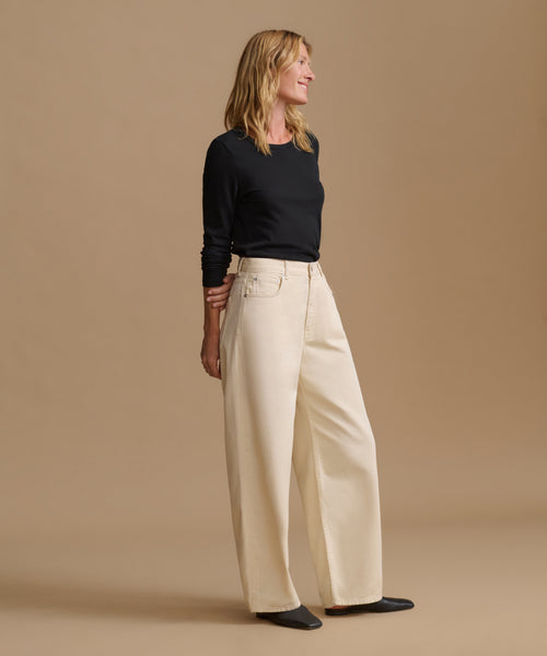 A woman modeling a black long-sleeve top and high-waisted, wide-leg cream pants against a solid beige studio background.