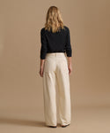 A woman viewed from behind wearing a black long-sleeve top and high-waisted, wide-leg cream trousers against a solid beige studio background.
