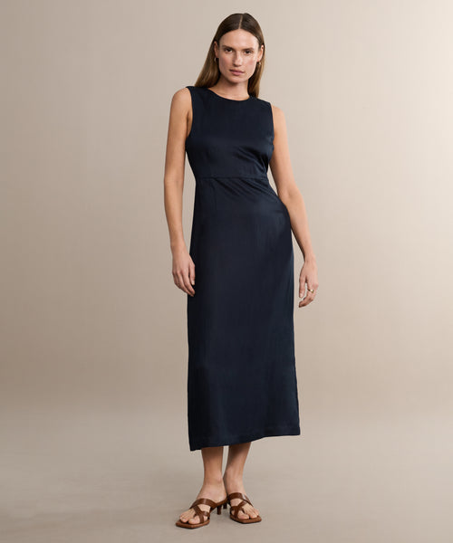 Against a plain light beige background, a woman with straight brown hair and minimal accessories wears the sleeveless Blair Dress in dark navy linen texture and brown flat sandals—a chic, neutral look ideal for spring.