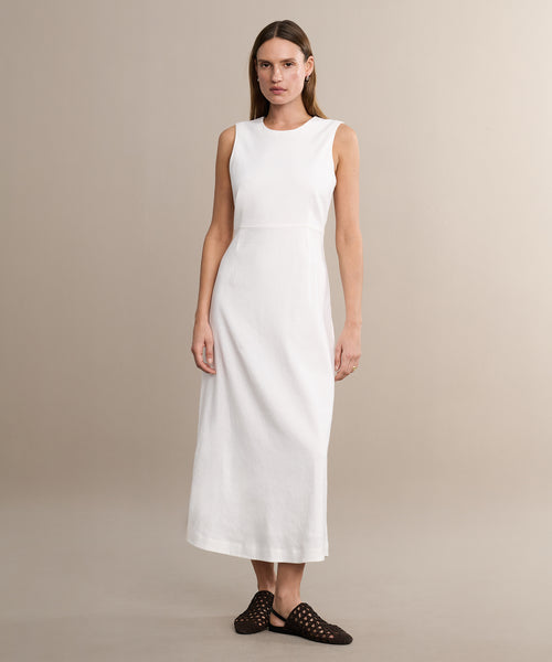A woman stands against a beige background wearing the sleeveless, ankle-length Blair Dress in white linen—an ideal spring piece. She pairs it with black woven flats, her straight brown hair framing her neutral expression.