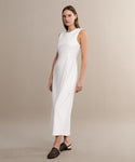 A woman in the Blair Dress—a sleeveless, ankle-length white linen dress—paired with black woven slip-on shoes stands against a beige background, looking to the side. The Blair Dress is a must-have for your spring wardrobe.