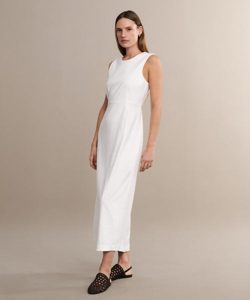 A woman in the Blair Dress—a sleeveless, ankle-length white linen dress—paired with black woven slip-on shoes stands against a beige background, looking to the side. The Blair Dress is a must-have for your spring wardrobe.