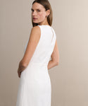 A woman with straight brown hair models the sleeveless Blair Dress in white, turning to glance over her shoulder. Its subtle linen texture makes it perfect for spring, showcased against a plain beige background.