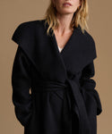 A close-up of a model wearing a black wool wrap coat with a wide shawl collar and matching waist tie, styled over a white top against a warm beige background.