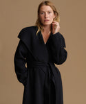 A woman wearing a black wrap coat with a tie waist belt, photographed against a solid warm beige studio background.