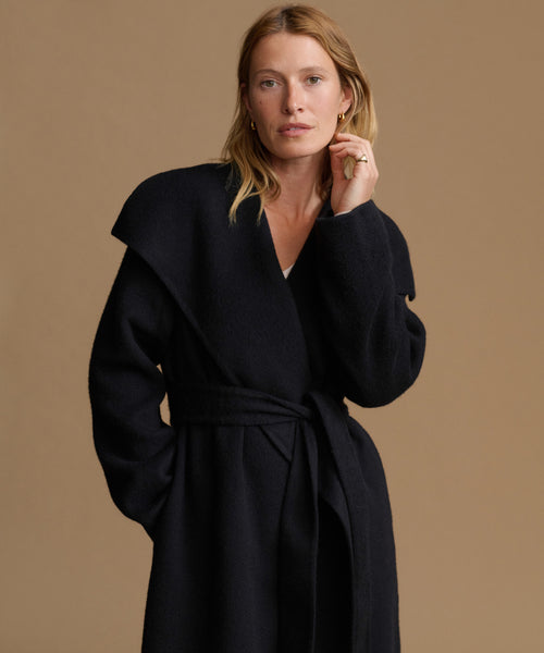 A woman wearing a black wrap coat with a tie waist belt, photographed against a solid warm beige studio background.
