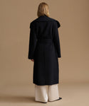 A model viewed from the back wearing a long black belted coat over wide-leg cream trousers, set against a solid warm beige studio background.