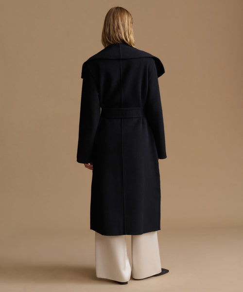 A model viewed from the back wearing a long black belted coat over wide-leg cream trousers, set against a solid warm beige studio background.